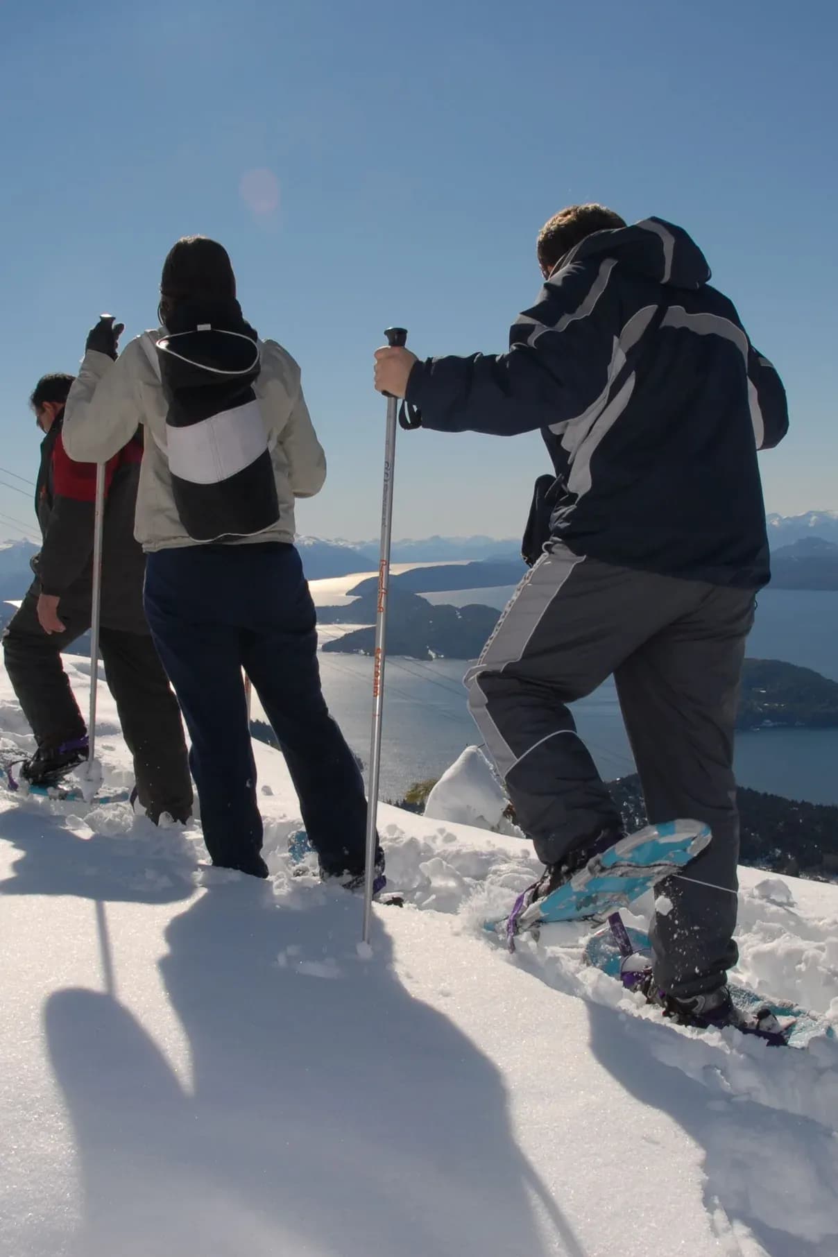 Tres personas de espaldas caminan con raquetas de nieve y bastones sobre una ladera nevada, contemplando una vista panorámica de un gran lago azul salpicado de islas y montañas lejanas bajo un cielo despejado.
