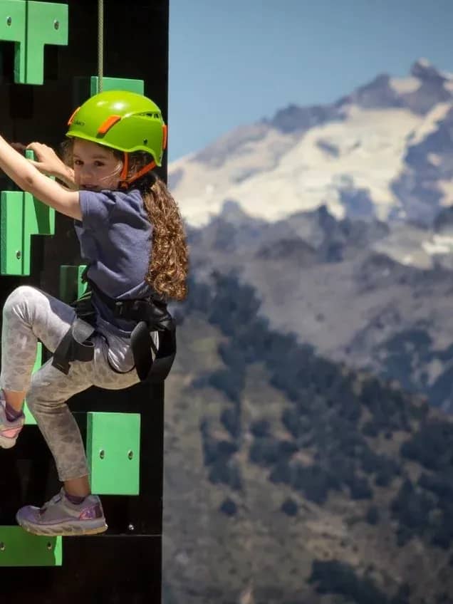 Niña con una casco verde y asegurada con una rnes y soga escalando una palestra con una montaña nevada de fondo